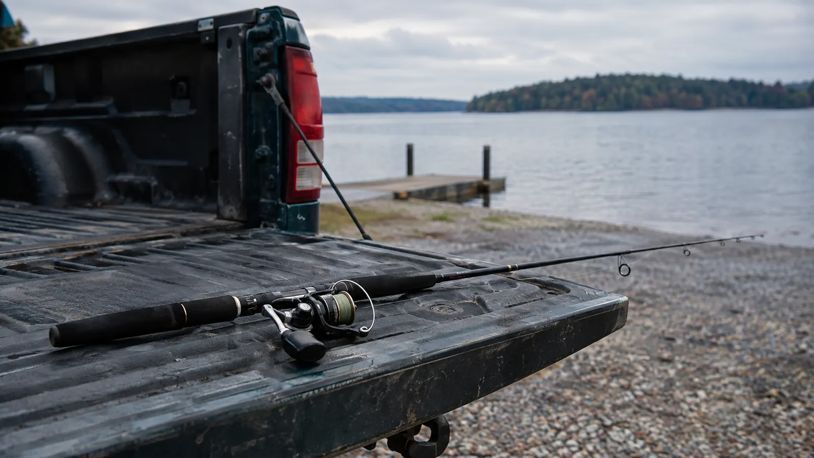 a spinning rod and reel lying across the tailgate of a pickup truck at a gravel boat ramp, with a lake and treeline behind