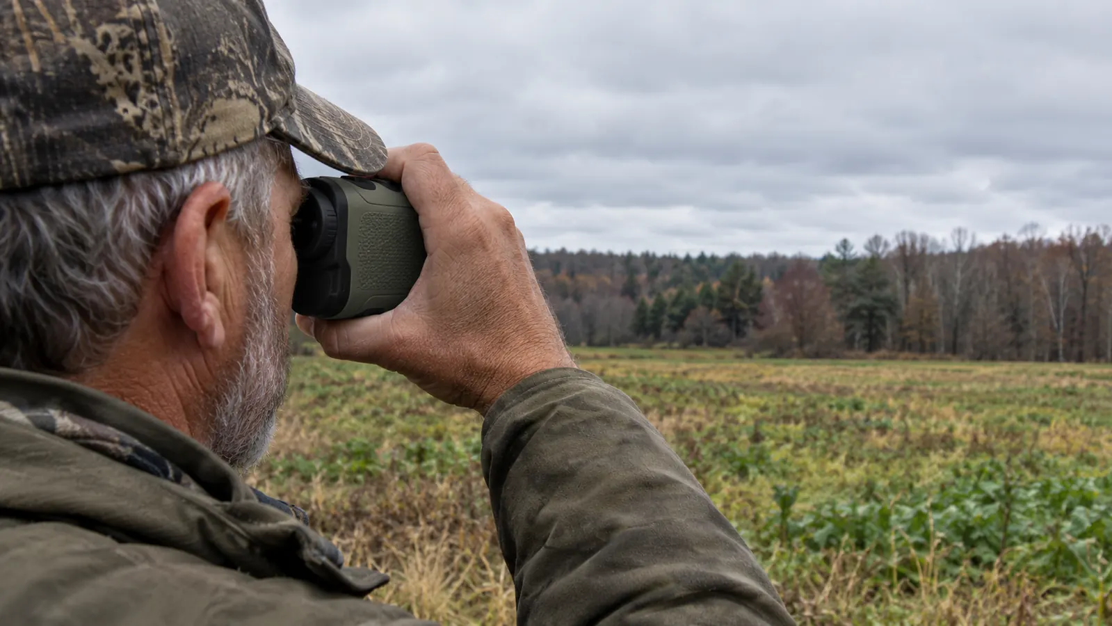 A hand holding a compact rangefinder up to one eye while looking across a food plot toward a treeline